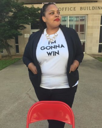 Tami Sawyer standing in front of a municipal office building, with a red folding chair propped up in front of her. Her hands are in her jacket pockets and she is wearing a T-shirt that says, "I'm gonna win."
