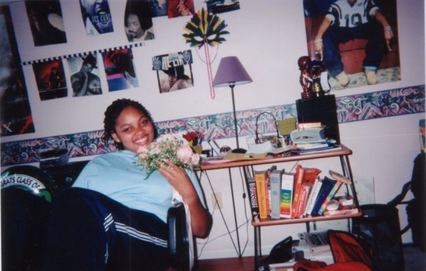 Sawyer as a teenager, sitting on a couch, holding flowers and smiling, with books and posters behind her.