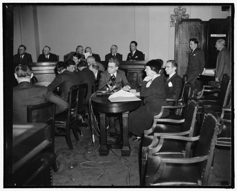 a black and white photo of a woman sitting at a desk surrounded by men above her at a dais